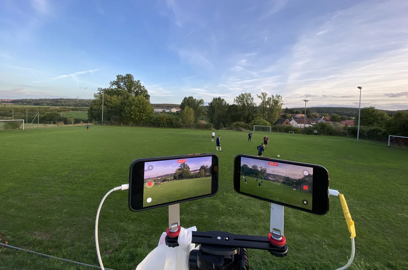 Zwei iPhones im Querformat auf einer Doppelschiene über einem Stativ am Rand eines Fußballplatzes, beide nehmen gerade auf.